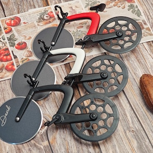 Peut inclure: Ensemble de trois coupe-pizzas en forme de vélos. Les coupe-pizzas ont des cadres noirs, blancs et rouges, et des lames circulaires. Les lames sont gravées du nom "David". Les coupe-pizzas sont présentés sur une surface en bois.
