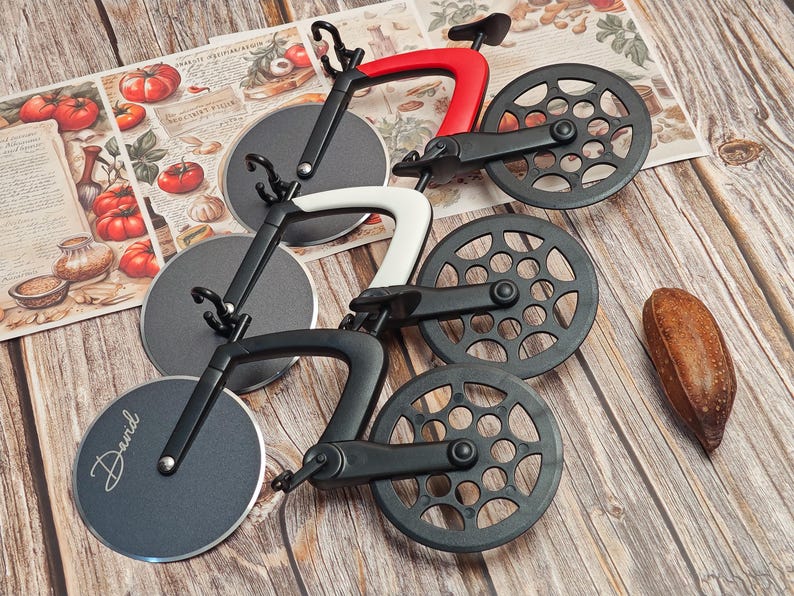 K&ouml;nnte beinhalten: Set mit drei Pizza-Schneidemessern in Form von Fahrr&auml;dern. Das Set enth&auml;lt Cutter mit schwarzen, wei&szlig;en und roten Rahmen und kreisf&ouml;rmigen Klingen. Die R&auml;der sind mit einem zahnradartigen Muster versehen. Auf einer Klinge ist der Name "David" eingraviert.