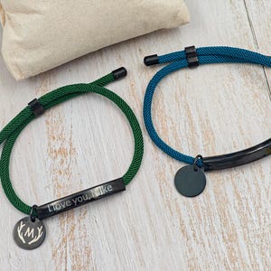May include: Two bracelets with dark green and teal rope bands. The green bracelet has a black rectangular plate engraved with "I love you, Mike" and a round charm with antlers and "MY". The teal bracelet has a black plate and a round charm.