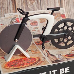 K&ouml;nnte beinhalten: Ein neuartiger Pizzaschneider in Form eines Fahrrads. Der Schneider hat einen schwarz-wei&szlig;en Rahmen, eine kreisf&ouml;rmige Klinge und einen schwarzen Sitz. Er liegt auf einer Pizza-Themenoberfl&auml;che mit einer Pizza-Illustration. Der Hintergrund ist eine Holzoberfl&auml;che.