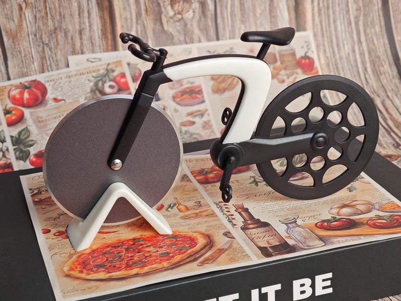 K&ouml;nnte beinhalten: Ein neuartiger Pizzaschneider in Form eines Fahrrads. Der Schneider hat einen schwarz-wei&szlig;en Rahmen, eine kreisf&ouml;rmige Klinge und einen schwarzen Sitz. Er liegt auf einer Pizza-Themenoberfl&auml;che mit einer Pizza-Illustration. Der Hintergrund ist eine Holzoberfl&auml;che.