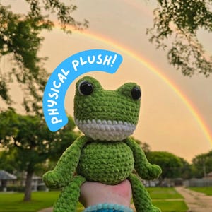 May include: A green crocheted frog plush with large black eyes and a white mouth area. A blue speech bubble graphic with the white text PHYSICAL PLUSH! is positioned above the frog. The frog has a textured yarn surface and long limbs.