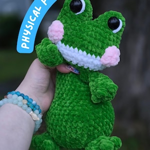 May include: A green crocheted frog plush toy featuring large black eyes with white accents, a wide white mouth, and round pink cheeks. A blue banner at the top left contains the white text PHYSICAL PLUSH! and a logo is in the bottom right corner.