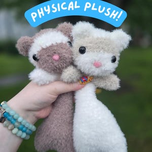 May include: Two fuzzy plush toys with rounded ears and small pink noses. One plush is brown with a white face patch, and the other is white with a light brown face patch. Both have black eyes and a blue banner above them reading PHYSICAL PLUSH!