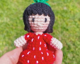 May include: A handmade crochet doll in the shape of a strawberry. The doll has a red dress with white spots, black hair, and green leaves on top. The doll is held in a hand against a blurred green background.