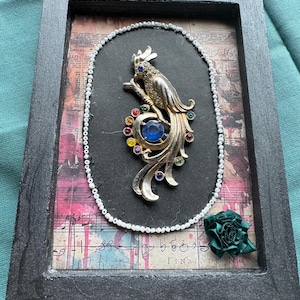 May include: A framed mixed-media art piece featuring a silver-toned parrot brooch with multicoloured rhinestones and a large blue centre stone. The brooch is encircled by a string of white beads on a background of vintage-style sheet music.