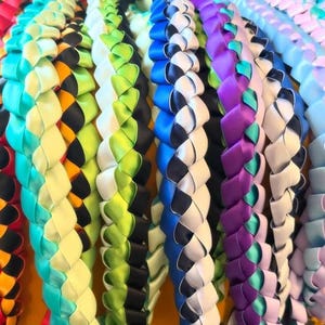 Peut inclure: Une collection de plusieurs colliers de rubans tressés disposés côte à côte. Chaque collier présente une combinaison de couleurs distincte, incluant des rubans de satin bleu vif, vert lime, violet, blanc, noir et turquoise tissés en motif.