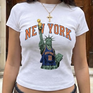 Puede incluir: Una camiseta blanca de manga corta con un gráfico de la Estatua de la Libertad vistiendo una camiseta de baloncesto azul y naranja con el número 11, sosteniendo un trofeo dorado, con el texto NEW YORK arqueado encima en letras naranjas y azules.
