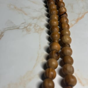 Op de afbeelding: Een gebedsketting gemaakt van lichtbruine houten kralen. De kralen zijn rond en geregen met een donkere draad. De kralen hebben een natuurlijke kleur en een gladde afwerking. De achtergrond is een wit en bruin marmerpatroon.