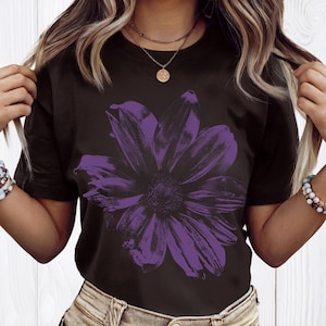 May include: Black t-shirt featuring a large, detailed purple flower graphic. The floral design is centered on the shirt, creating a bold contrast against the dark background. The shirt is a classic crew neck style.