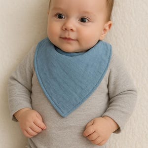 Puede incluir: Un bebé con un babero azul claro sobre un body gris de manga larga. El babero es triangular y está hecho de tela suave. El bebé está acostado sobre una superficie blanca.