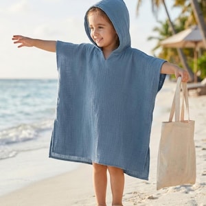 May include: A young child wearing a blue hooded poncho stands on a sandy beach, arms outstretched. The poncho is made of a textured fabric. The child is also wearing sandals and holding a beige tote bag.