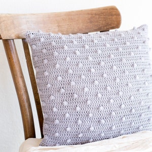 May include: A light gray, rectangular crochet pillow with a textured, dotted pattern. The pillow is resting on a wooden chair, showcasing its handmade quality and soft texture. The pillow's neutral color complements various home decor styles.