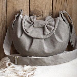 May include: A light gray corduroy shoulder bag with a large bow detail on the front. The bag has a long, adjustable strap and a metal buckle. The bag is sitting on a white blanket against a wooden background.