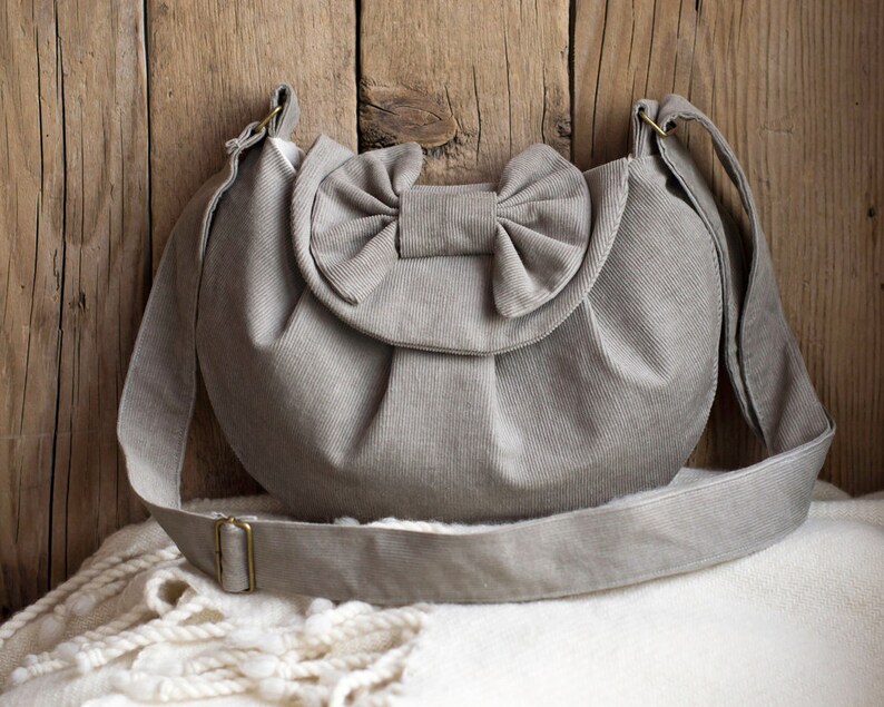 May include: A light gray corduroy shoulder bag with a large bow detail on the front. The bag has a long, adjustable strap and a metal buckle. The bag is sitting on a white blanket against a wooden background.