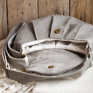 May include: Open view of a gray corduroy shoulder bag with a flap closure and a long adjustable strap. The bag has a magnetic snap closure and brass-colored hardware. The interior is lined with white fabric.