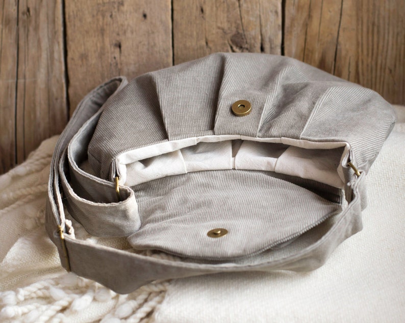 May include: Open view of a gray corduroy shoulder bag with a flap closure and a long adjustable strap. The bag has a magnetic snap closure and brass-colored hardware. The interior is lined with white fabric.
