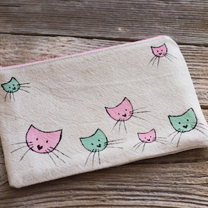 May include: A rectangular, beige canvas pouch with a pink zipper and a pink lining. The pouch is decorated with multiple cartoon cat faces in pink and mint green, each with black whiskers. The pouch is lying on a wooden surface.