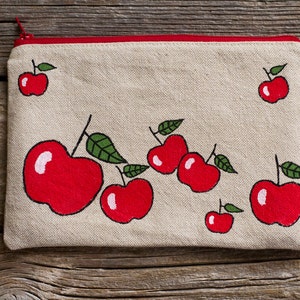 May include: A rectangular beige canvas pouch with a red zipper and trim. The pouch is decorated with hand-painted red apples with green leaves. The apples are of varying sizes and are scattered across the surface of the pouch.