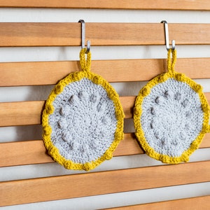 May include: Two gray and yellow crocheted potholders hanging on a wooden slat wall. The potholders are round with a gray center and a yellow border. Each potholder has a loop for hanging.