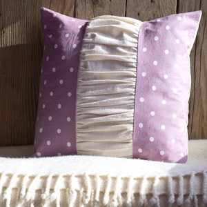 May include: A decorative pillow with a lavender polka dot pattern on the sides and a cream-colored, ruffled center. The pillow is square and rests on a white, fringed blanket against a wooden background.