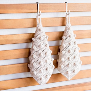 May include: Two off-white, handmade crochet washcloths with a textured, bumpy design. The washcloths are hanging on a wooden slatted wall with silver hooks. The crochet work has a unique, organic appearance.