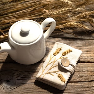 May include: A white ceramic teapot with a lid and a curved handle sits on a rustic wooden surface. Beside it is a beige linen pouch with embroidered wheat stalks and a wooden button closure. Dried wheat stalks are in the background.