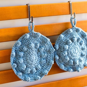 Puede incluir: Dos salvamanteles redondos de ganchillo azul claro con un patrón de puntos en relieve texturizado. Cada salvamanteles tiene un lazo para colgar y está suspendido de un gancho de metal. Los salvamanteles se muestran sobre un fondo de listones de madera.