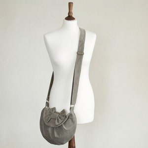 May include: A gray suede crossbody bag with a bow detail on the front flap. The bag has a long, adjustable strap and a metal buckle. The bag is displayed on a white mannequin.