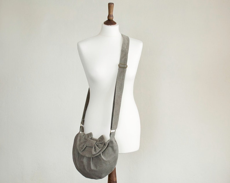 May include: A gray suede crossbody bag with a bow detail on the front flap. The bag has a long, adjustable strap and a metal buckle. The bag is displayed on a white mannequin.