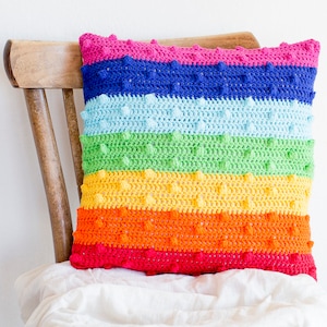 May include: A handmade, square crochet pillow with a rainbow color scheme. The pillow features horizontal stripes of red, orange, yellow, green, light blue, dark blue, and pink. Each stripe has a textured, bobble stitch design. The pillow is resting on a wooden chair.