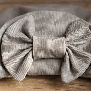 May include: A close-up of a light gray corduroy bow. The bow is the focal point, with a detailed texture and a prominent knot in the center. The fabric has a ribbed pattern, and the bow is set against a matching background.