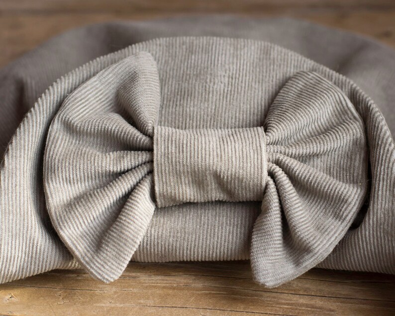 May include: A close-up of a light gray corduroy bow. The bow is the focal point, with a detailed texture and a prominent knot in the center. The fabric has a ribbed pattern, and the bow is set against a matching background.