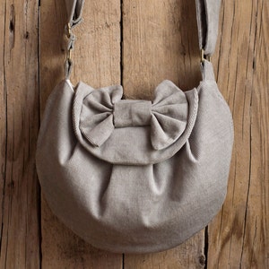 May include: A light gray corduroy shoulder bag with a large bow detail on the front flap. The bag has a rounded shape and a long, adjustable strap. The bag is made of a soft, textured fabric and has gold-colored hardware.