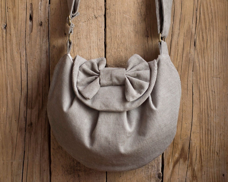 May include: A light gray corduroy shoulder bag with a large bow detail on the front flap. The bag has a rounded shape and a long, adjustable strap. The bag is made of a soft, textured fabric and has gold-colored hardware.