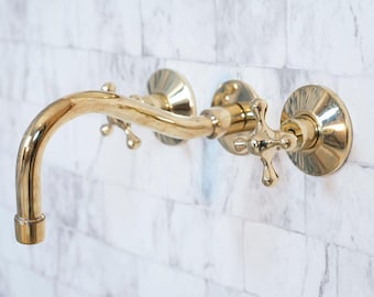 May include: A wall-mounted, gold-colored faucet with a curved spout and cross-shaped handles. The faucet is made of brass and has a polished finish. The fixture is mounted on a white brick wall, adding a touch of vintage elegance to any bathroom.
