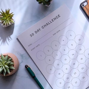 May include: A 30-day challenge tracker with numbered circles, a pen, and a phone with a wooden case. The tracker has spaces for goal, start date, and end date. Two small potted succulents are also in the image.