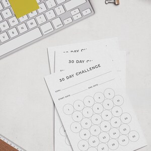 May include: A top-down view of a workspace with a white keyboard, sticky notes, and a pen. Several white paper sheets with the text "30 DAY CHALLENGE" are visible, featuring numbered circles for tracking progress. A ruler and a wire basket are also present.