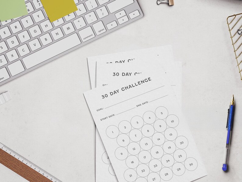 May include: A top-down view of a workspace with a white keyboard, sticky notes, and a pen. Several white paper sheets with the text "30 DAY CHALLENGE" are visible, featuring numbered circles for tracking progress. A ruler and a wire basket are also present.