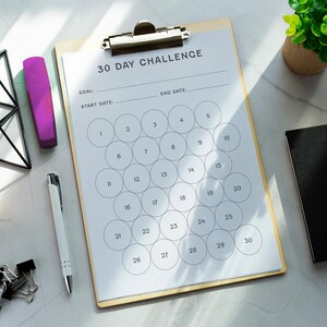 May include: A clipboard with a 30-day challenge checklist, a pen, a purple highlighter, a yellow highlighter, a black notebook, and binder clips are arranged on a marble surface. A small potted plant and a geometric wire frame are also present.