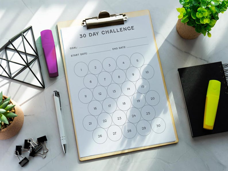 May include: A clipboard with a 30-day challenge checklist, a pen, a purple highlighter, a yellow highlighter, a black notebook, and binder clips are arranged on a marble surface. A small potted plant and a geometric wire frame are also present.