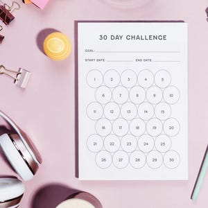 May include: A white 30-day challenge tracker with circles numbered 1-30. The text "30 DAY CHALLENGE" is at the top. The image includes headphones, a pen, and office supplies on a pink surface.