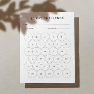 May include: A white 30-day challenge tracker with the text "30 DAY CHALLENGE" at the top. It includes spaces for a goal, start date, and end date, along with 30 numbered circles for tracking progress. The tracker is on a brown surface.