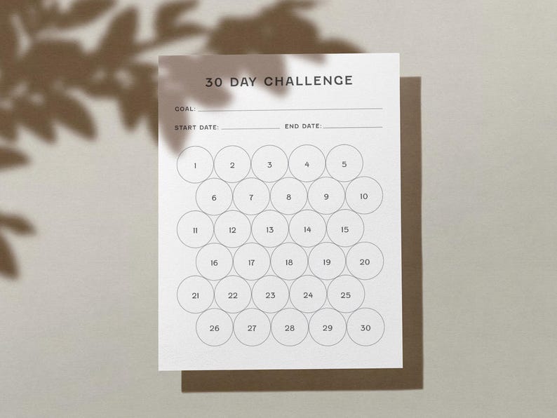 May include: A white 30-day challenge tracker with the text "30 DAY CHALLENGE" at the top. It includes spaces for a goal, start date, and end date, along with 30 numbered circles for tracking progress. The tracker is on a brown surface.