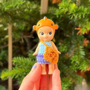 May include: A small figurine wearing an orange clownfish headpiece, a light blue crocheted top, and a purple crocheted bottom. The figurine carries a small, textured orange crocheted bag across its body, featuring a round shape with dark brown accents.