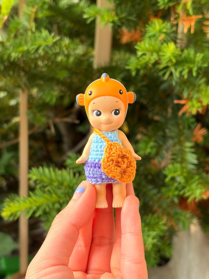 May include: A small figurine wearing an orange clownfish headpiece, a light blue crocheted top, and a purple crocheted bottom. The figurine carries a small, textured orange crocheted bag across its body, featuring a round shape with dark brown accents.