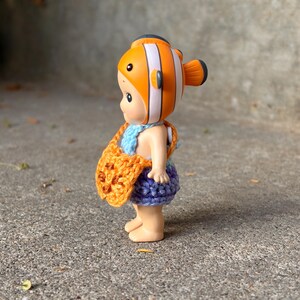May include: A small figurine wearing a clownfish-themed headpiece with orange, white, and black stripes. The figure is dressed in a light blue crocheted top, a purple crocheted skirt, and carries a small orange crocheted bag with dark brown accents.