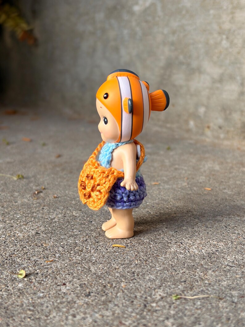 May include: A small figurine wearing a clownfish-themed headpiece with orange, white, and black stripes. The figure is dressed in a light blue crocheted top, a purple crocheted skirt, and carries a small orange crocheted bag with dark brown accents.