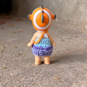 May include: A small figurine wearing a crocheted dress with a blue and purple gradient pattern. The figure has a round orange and white headpiece with a fin-like detail, small white wings on the back, and a thin orange strap across the shoulder.