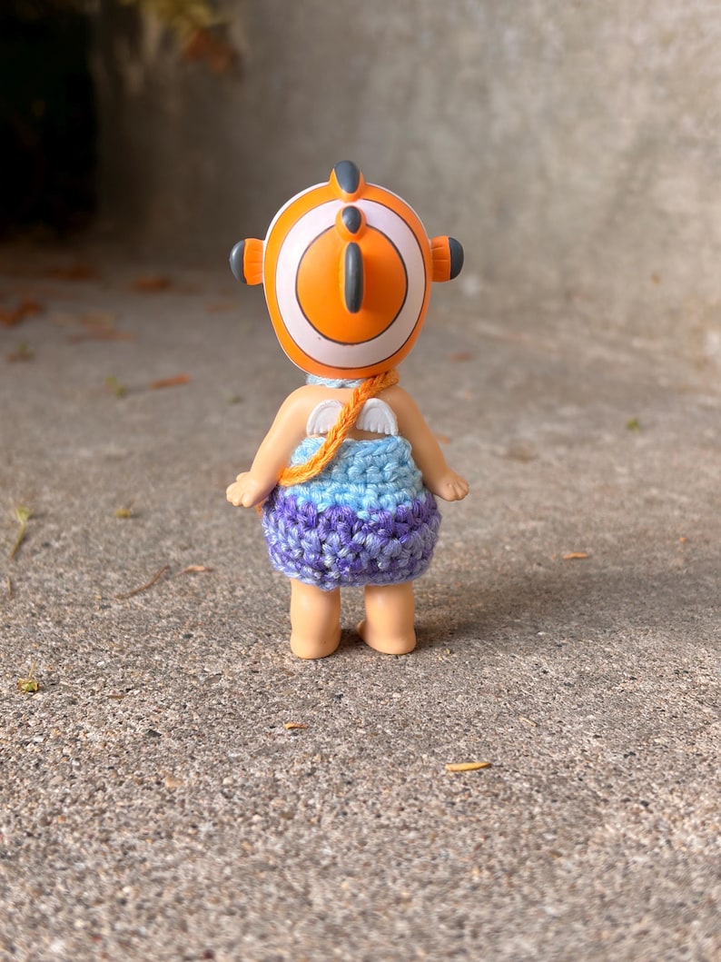 May include: A small figurine wearing a crocheted dress with a blue and purple gradient pattern. The figure has a round orange and white headpiece with a fin-like detail, small white wings on the back, and a thin orange strap across the shoulder.